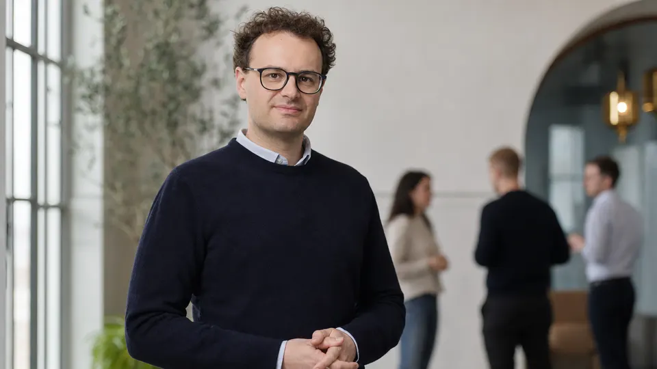 A man in three-quarter view stands in a bright modern office with colleagues blurred behind him.