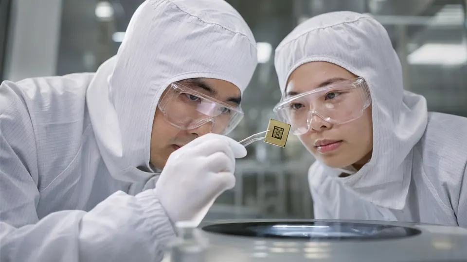 Two engineers in clean-room suits examine a small processor package at a lab bench under bright light.