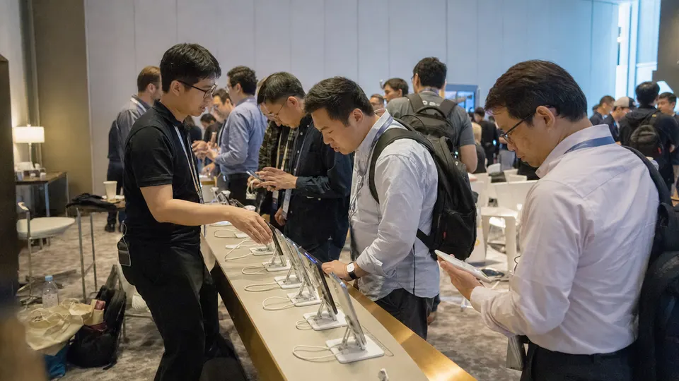 Visitors at a convention hall demo table examine smartphones on stands while a technician explains one of them, with gold trim along the table edge.