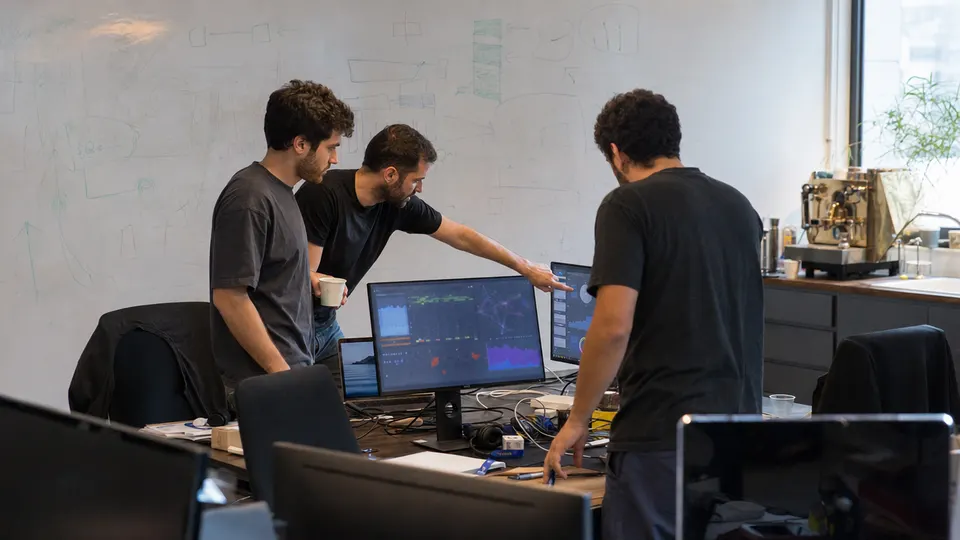 Three engineers gather around a desk with two monitors displaying dashboards in a Tel Aviv startup office, with a brass espresso machine on a counter behind them.