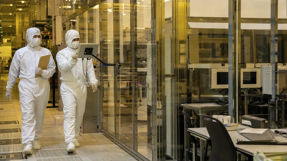 Two technicians in white cleanroom suits walk through a chip fabrication plant corridor lit in amber, past glass-walled equipment bays.