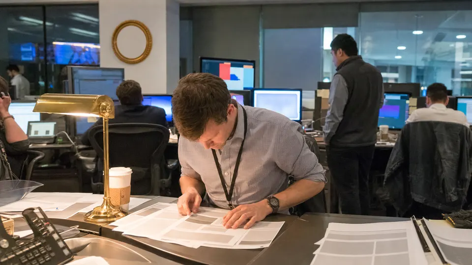 A producer in shirtsleeves reviews printed story pages at a newsroom desk lit by a brass lamp, with market charts on blurred monitors behind.