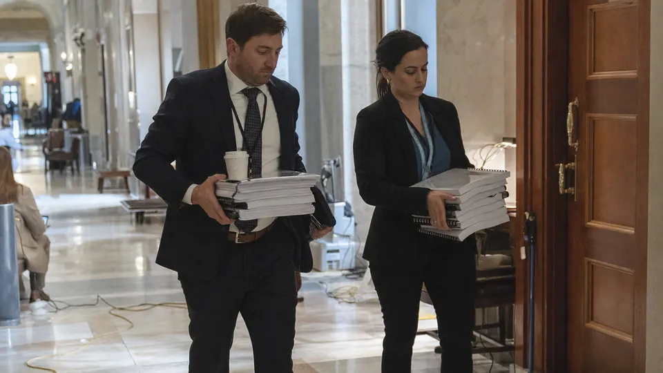 Two congressional staffers carry stacks of bound reports down a marble hallway in a Senate office building.