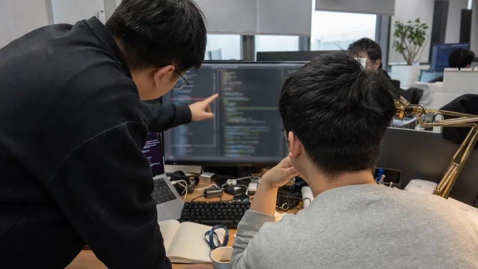 Two software engineers review code on a monitor at a shared desk in an office, with a brass lamp and a cup of tea beside the keyboard.