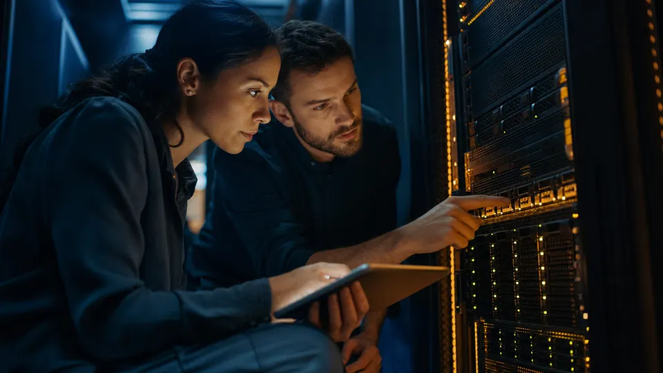 Two security engineers examine a server rack in a dimly lit data center, lit by blue and amber indicator lights.