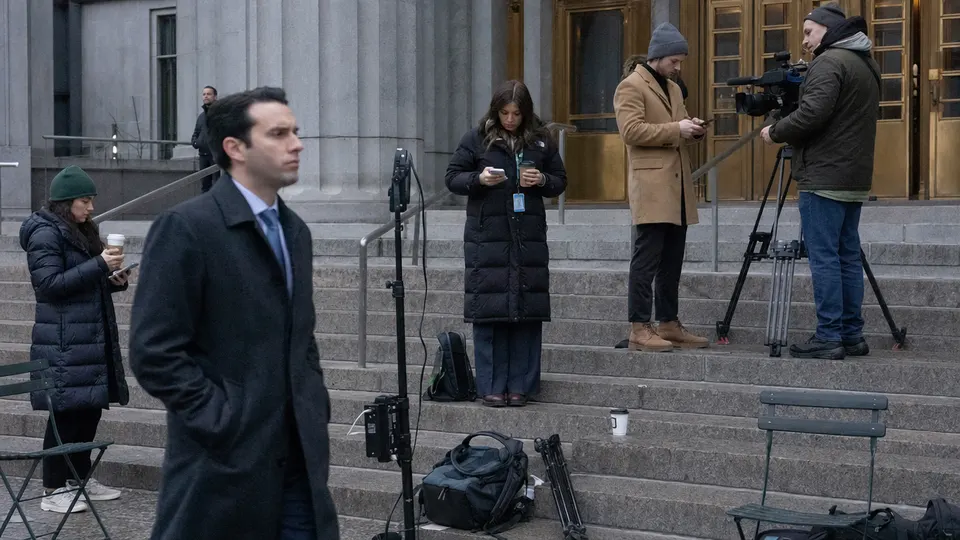 Reporters and a camera operator wait on the steps of a federal courthouse in Manhattan as a man in a dark overcoat walks past, brass door fittings gleaming.