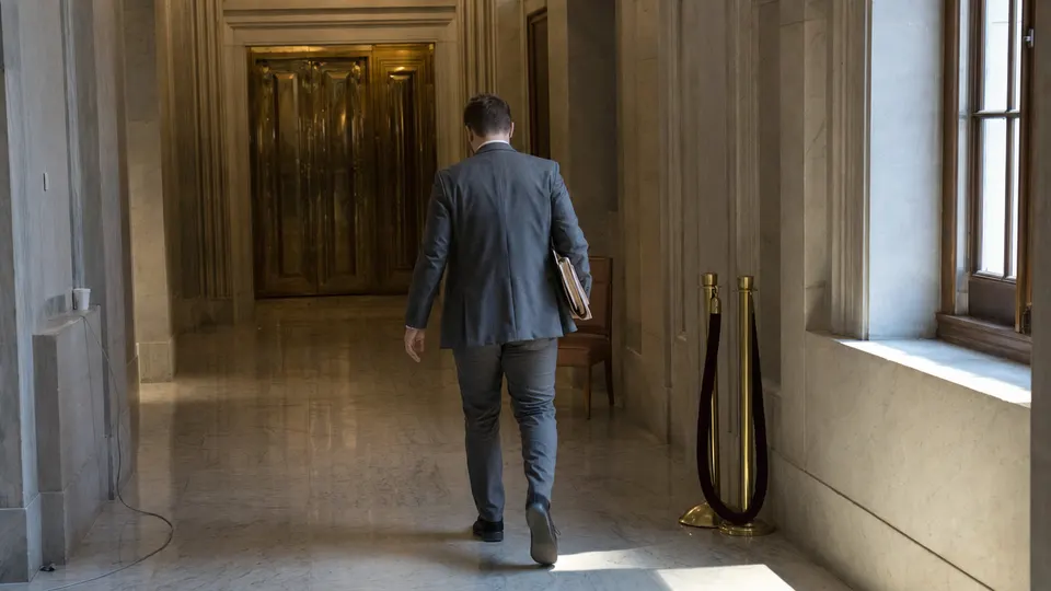 A congressional staffer carries a folder down an empty marble corridor of a Senate office building, with brass elevator doors at the far end.