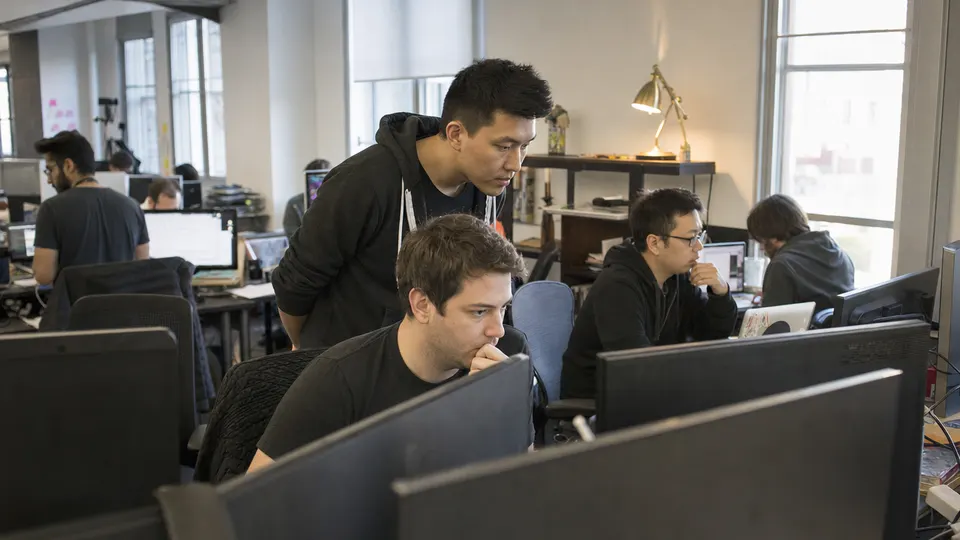 Engineers work at shared desks in a sunlit San Francisco startup office while a colleague leans over a monitor beside a brass desk lamp.