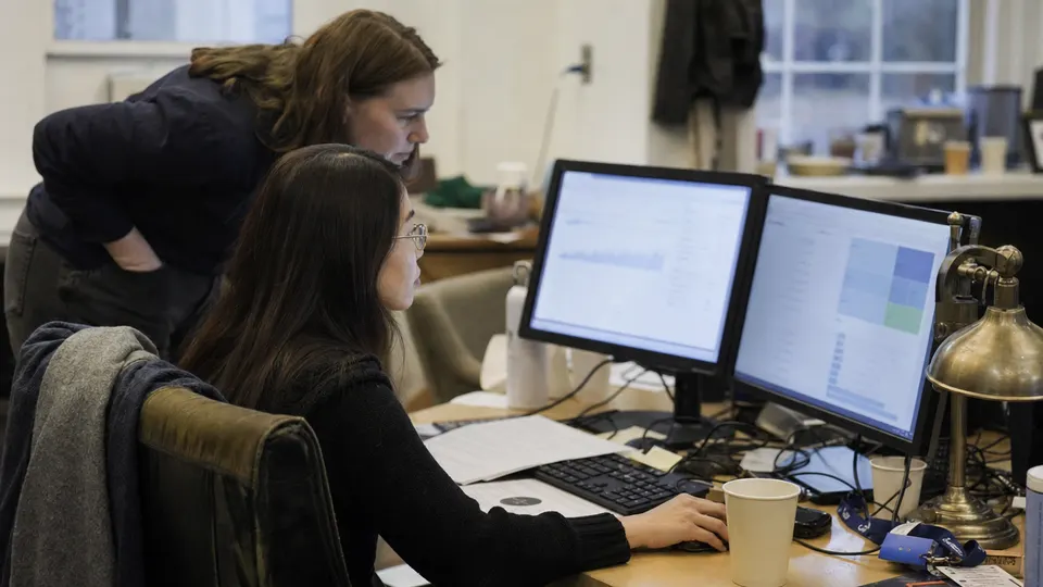 Two Wikimedia staff members at a cluttered office desk look at monitors showing graphs, with a brass desk lamp in the afternoon light.