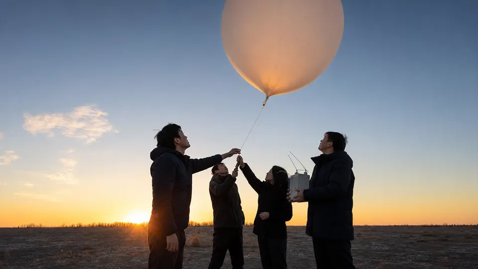 Engineers launching a large weather balloon at sunrise in an open field, watching it rise into the sky.