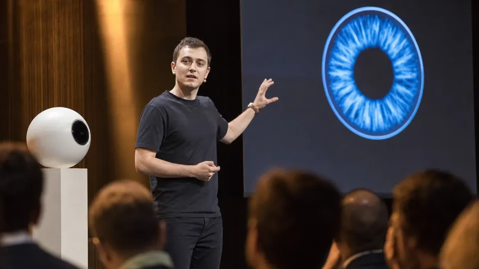 Illustration-style news photo of a World co-founder presenting on a conference stage next to a white iris-scanning orb, with a blue badge graphic on a screen behind him.