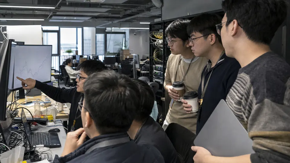 Engineers in a Beijing office gathered around a monitor showing a rising line chart, with server racks behind them.