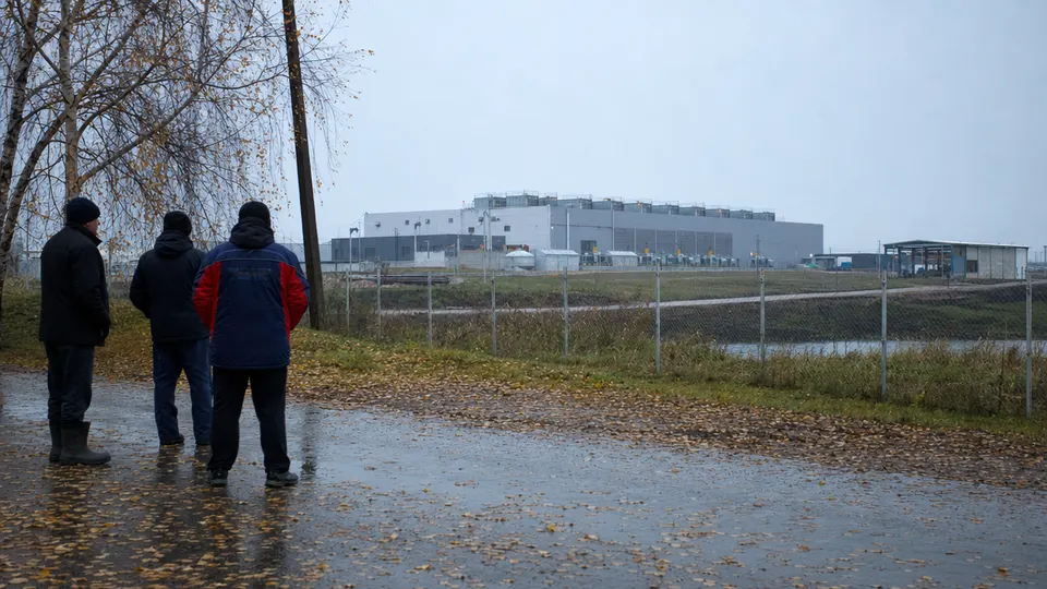 Residents stand beside a roadside fence near an industrial complex in Sasovo, Russia, in early morning light, with golden birch leaves on the pavement.