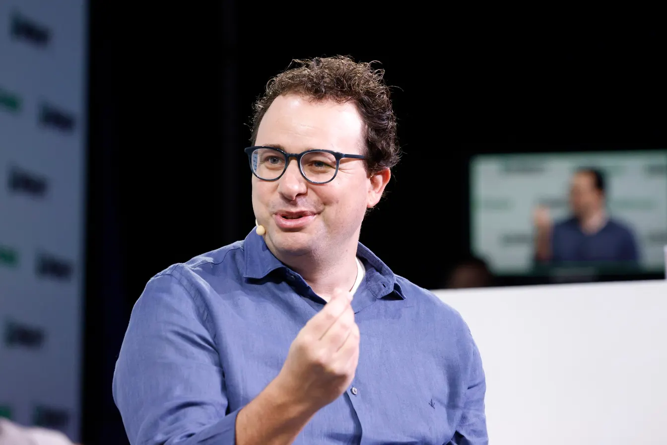 Dario Amodei speaking on stage at TechCrunch Disrupt 2023, in a blue shirt and glasses, gesturing as he talks